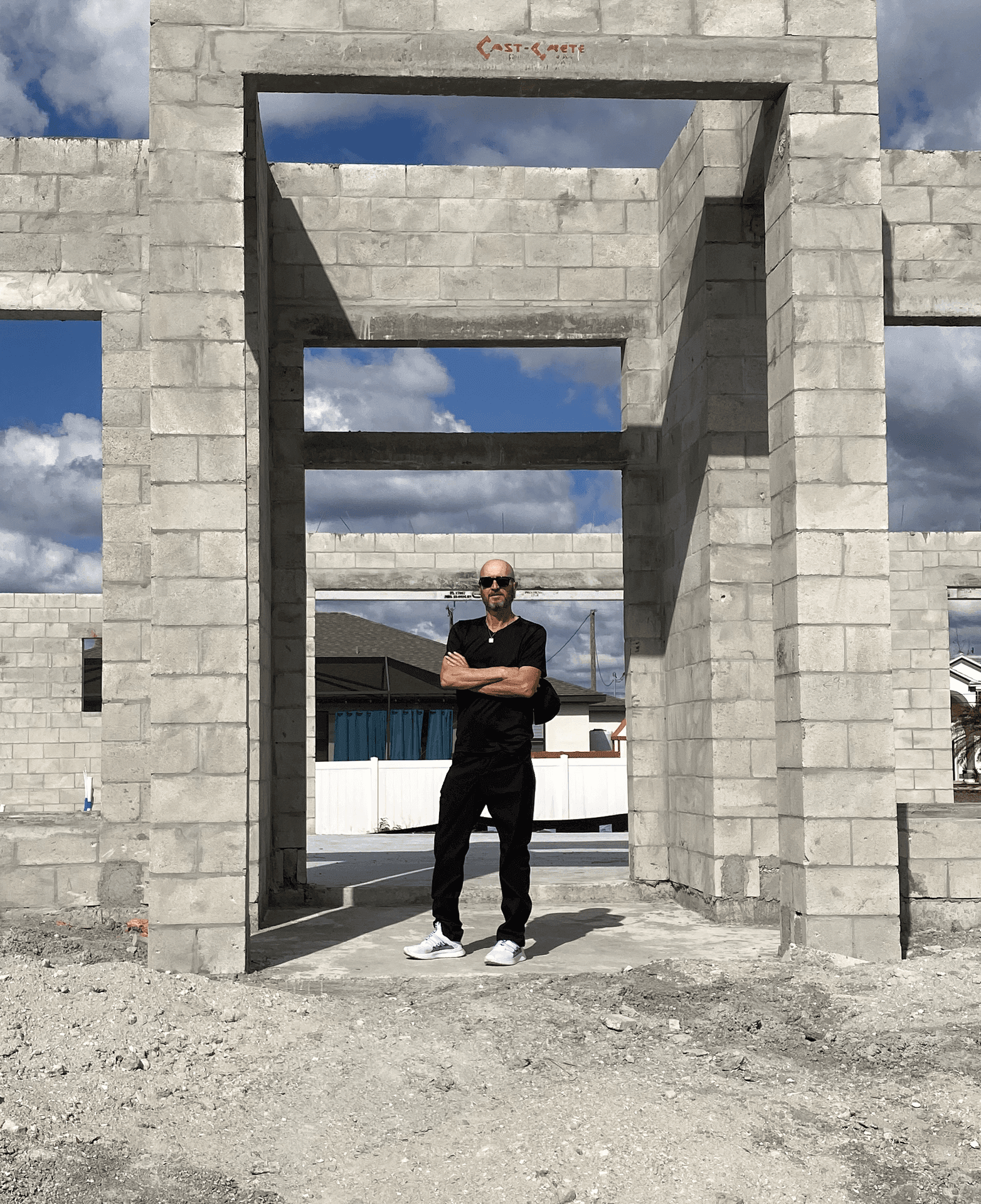 Man standing with arms crossed inside an unfinished concrete structure with blue sky and clouds in the background.